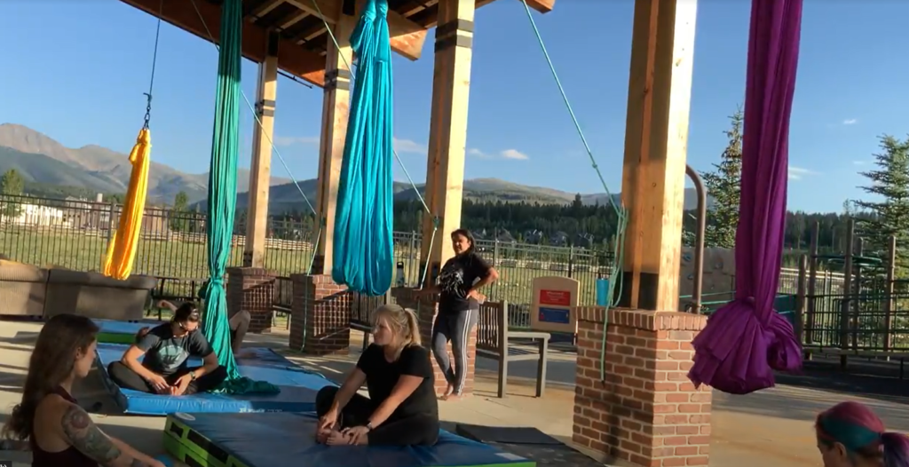 An outdoor aerial silks class is in session under a wooden shelter with a mountainous backdrop bathed in golden sunlight. A group of people is gathered around, with some sitting on mats on the floor, preparing for or resting between exercises. 