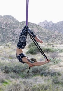 A person is performing an aerial maneuver on a hoop suspended in the air, with a natural, rocky landscape in the background. 