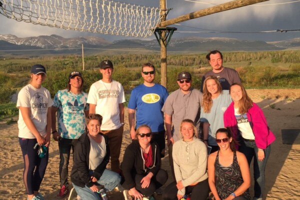 Group photo of a volleyball team on a sand court with a net overhead and a scenic backdrop of mountains and clouds.