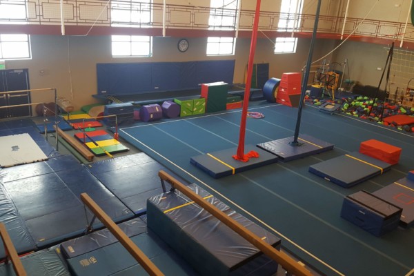 Indoor gymnastics training area with various equipment such as mats, beams, and padded blocks. The space is well-lit with a high ceiling and a large clock on the wall, suggesting a professional or school gymnasium setting for gymnastics practice.